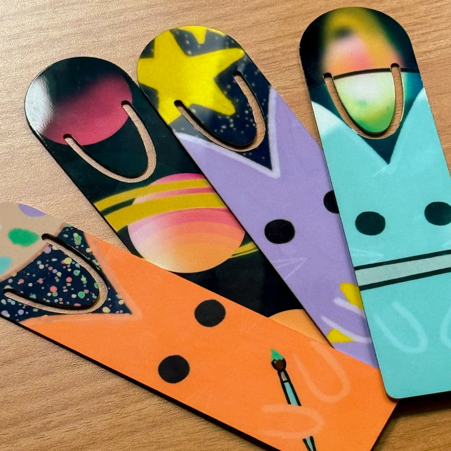 Four colorful, abstract bookmarks with playful designs: a cosmic cat face, a starry night with a rocket, a polka-dot and moon pattern, and a vibrant geometric sun—each with a gold U-shaped slot for securing pages.